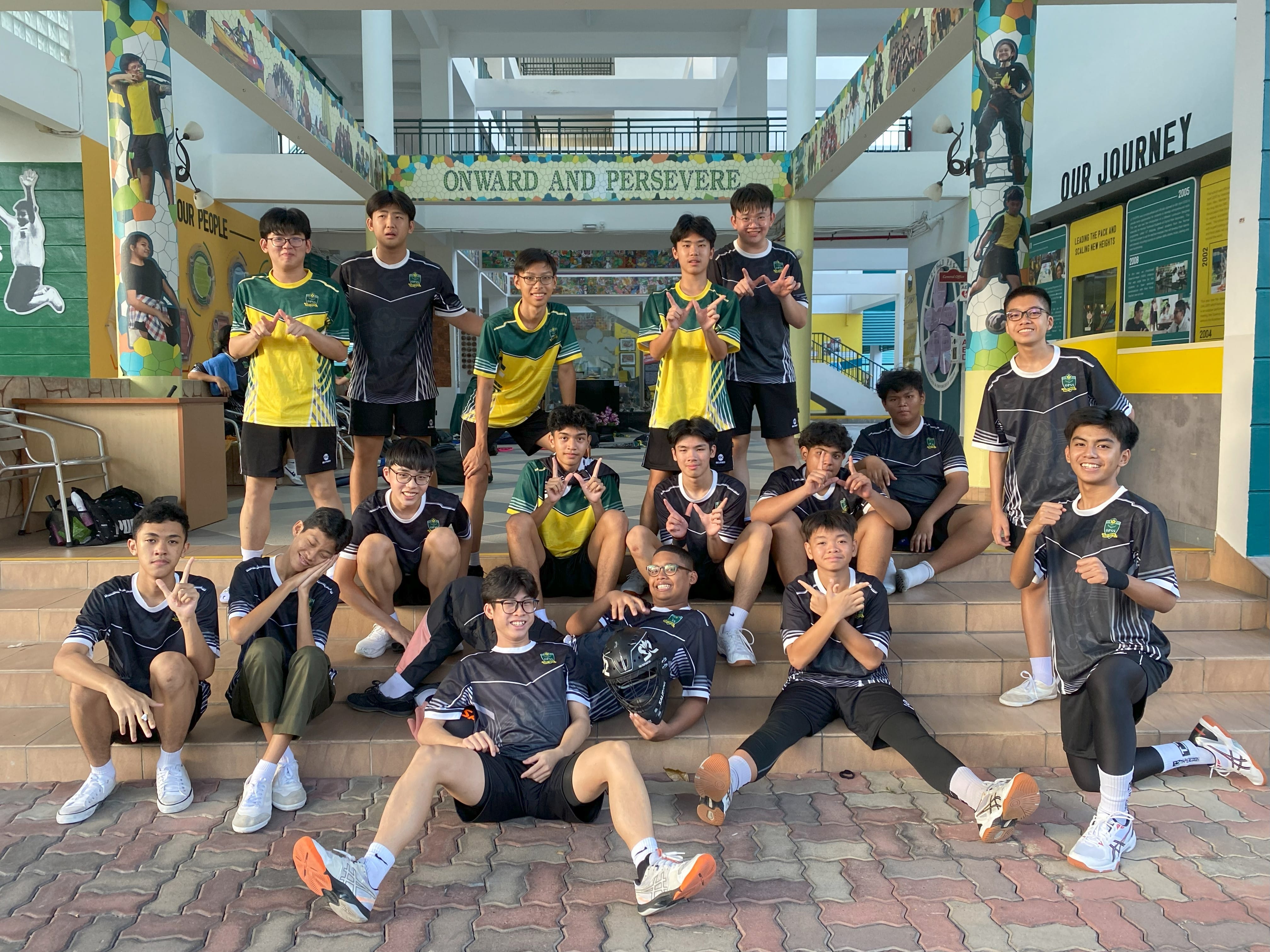A school boys' floorball team posing playfully on outdoor steps beneath a walkway banner that reads "ONWARD AND PERSEVERE". The players are wearing dark grey, green, and yellow sports jerseys with black shorts, smiling and making hand gestures.